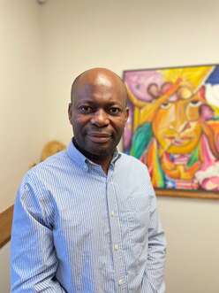 A man in a striped button down shirt stands smiling in the foreground, with a blurred abstract painting behind him. 