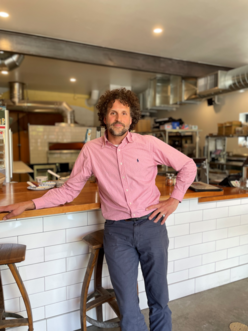A man in a pink dress shirt stands in front of a food counter.