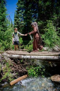 Outoors, background of pine trees and blue sky, foreground of the bare log bridge crossing over a running creek; a male volunteer extends his hand to support a female participant to cross the bridge. 