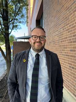 Danny Beus (glasses, beard and mustache)  stands in front of the IRC office in a suit and tie smiling at the camera. Primarily a brick wall background with a cropped tree and blue sky.