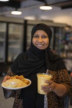 Chef Najati holds a plate of food and a drink while dressed in a black headscarf.