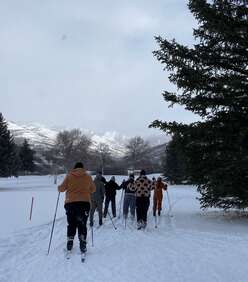 A group of youth ski away from the camera, with a large tree in the foreground right and trees and mountains in the distance. 