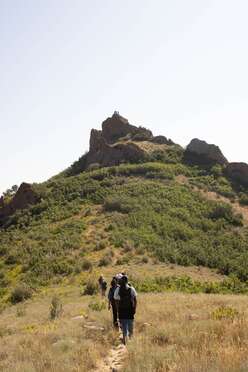 A group of refugee youth hike along a dirt trail headed toward the peak of a brush-covered mountain ahead. Along the horizon, figures can be seen standing on the mountaintop.