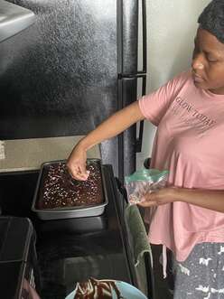 Refugee woman stands in front of her oven stovetop in a pink shirt adding the finishing touches (sprinkles!) to a chocoloate cake in a rectangle cake pan.