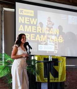 Carolina Terrazas, dressed in a white top and white pants, speaks into a standing microphone with a leafy plant, an IRC-logo flag, and a projector displaying the event title: New American Dream Lab.