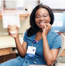 Torbertha Torbor poses sitting down in her scrubs. She is holding a cup of coffee and smiling.