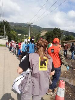An IRC staff member stands with her back to the camera. In front of her is a line of people waiting to receive supplies.