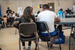 In the IRC's Welcome Center for asylum seekers, a husband and wife sit with their back to the camera as they discuss their journey as asylum seekers. The wife is holding their 2-year-old daughter. 