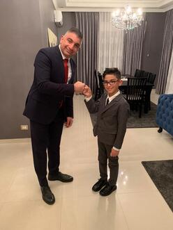 Maha's eldest son and 13-year-old grandson, both wearing suits and ties, fist bump and smile for the camera.