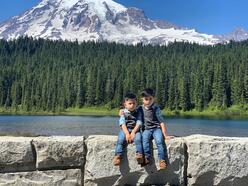 Two young boys sit on a stone wall in front of a lake and mountain.