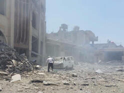 A man walks toward a crushed car in front of a recently bombed building. There is rubble and dust and smoke in the air.