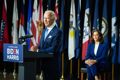 President Joe Biden stands at a podium giving a speech while Vice President Kamala Harris sits on stage. The new administration takes office after four years of Trump Administration policies that wreaked havoc on the lives of refugees and asylum seekers.