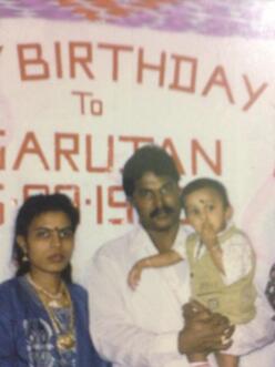 Shanthi and her  husband Sivakumar, who is holding toddler Sarujen, stand in front of a sign that says Happy Birthday to Sarujen