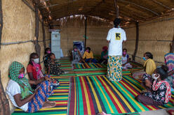 An IRC staff member wearing an IRC logo t-shirt speaks with children and adults sitting on mats on the floor in an IRC safe space for Tigrayan refugees in Sudan.