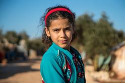 Heba, a ten-year-old Syrian girl stands with a serious expression in the dusty street of the displacement camp where her family lives.