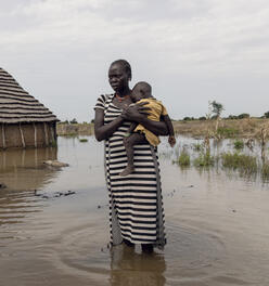 With water above her ankles, a mother holds her child and stands in front of her flooded home.