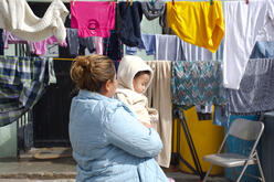 A woman wearing a blue winter coat looks away from the camera while holding a toddler. Laundry dries on clotheslines behind her.
