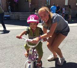 A woman IRC volunteer leans to hold the handlebars of the pink and white bike of a little girl as she is riding the bike wearing a bring pink helmet, both smile for the camera.