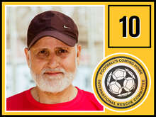 Ein älterer Mann mit weißem Bart und brauner Baseballkappe lächelt leicht in die Kamera und trägt ein rotes T-Shirt.