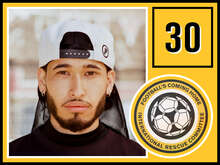 Ein junger Mann mit Bart und Ohrringen schaut ernst in die Kamera, trägt eine weiße Baseballkappe rückwärts.