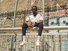 Ein Mann in einem weißen Hemd und dunklen Hosen sitzt auf einem Zaun vor einer mit Graffiti bedeckten Wand.