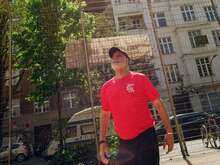 Ein Mann in einem roten T-Shirt und schwarzer Kappe schaut bei hellem Sonnenschein in die Luft, während ein Ball über ihm schwebt; im Hintergrund sind ein Zaun und mehrstöckige Gebäude zu sehen.