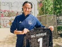 Ein Mann im SC Horus Trainingsanzug hält stolz ein Trikot mit der Nummer 1 vor einem mit Graffiti bedeckten Gebäude hoch.