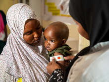 한 어머니가 국제구조위원회 영양담당 직원이 영양실조 징후를 검사하기 위해 자녀를 안고 뮤악테이프로 팔 둘레를 측정하는 것을 돕고 있습니다.