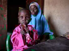 En liten flicka håller en nötkräm för behandling mot undernäring i sina händer. Hennes mamma sitter bakom henne.