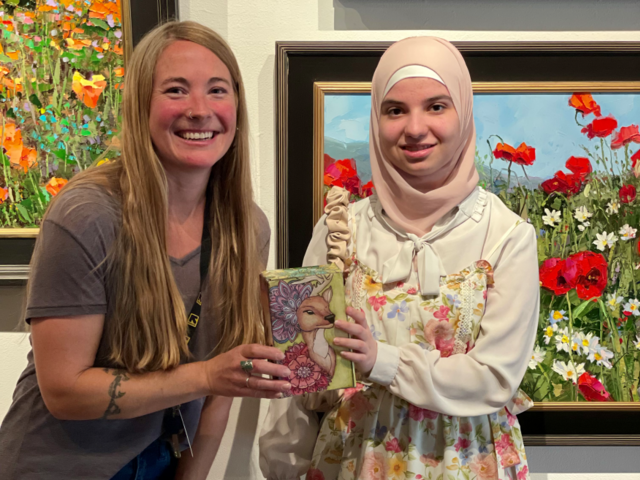 Two women stand alongside each other holding a small box with a drawing of a deer and mandala