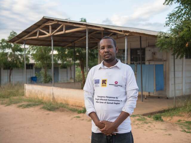 Ein Mann steht lächelnd vor einem kleinen, einfachen Gebäude mit Wellblechdach in einer ländlichen Gegend.