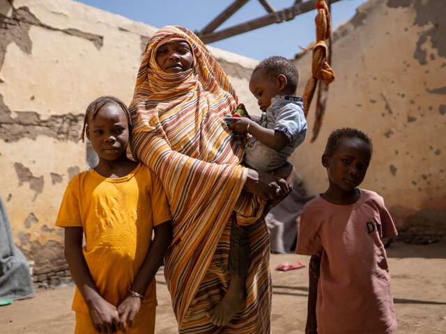 En mamma håller sitt yngsta barn i famnen. Två andra barn står vid sidan om henne i en skyddsbostad i Sudan.