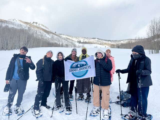 8 participants dressed in snow gear wearing snowshoes stand side by side in front of a snowy landscape with snow-covered mountains in the background and a cloudy sky. The central figures hold a flag with a while field, blue logo noting "Summit Journeys".