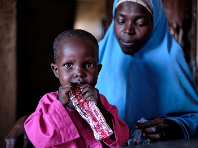 En liten pojke sitter i sin mammas knä och håller i en nötkrämspåse, han får behandling mot undernäring i ett flyktingläger i Nigeria.