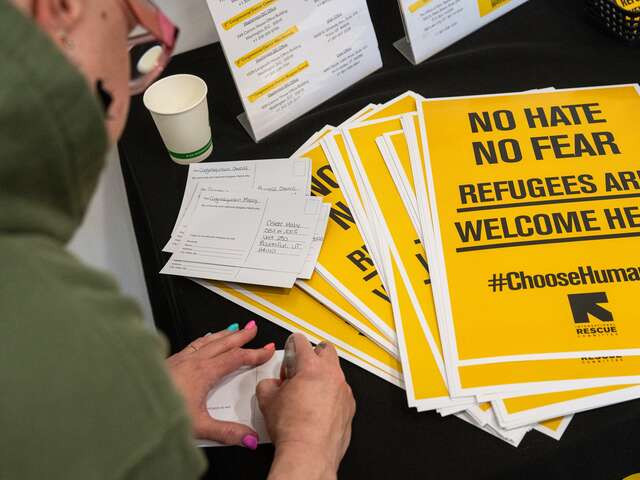 The photographer peeks over the shoulder of a woman in a green hoodie writing a postcard to her elected officials. She's writing her postcard alongside a pile of posters with a call to action: "No Hate, No Fear, Refugees are welcome here!"