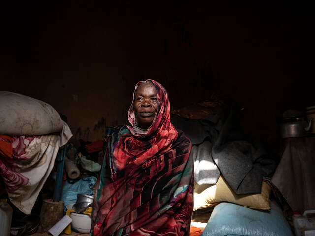 En äldre kvinna i Tchad står i ett mörkt tält, solen lyser in på hennes ansikte.