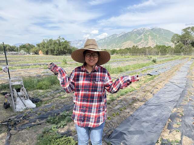 Hser Nay stands centered in the photo in a large straw hat, red plaid long-sleeve shirt, and jeans, smiling with her arms up showing off the farm & scenic mountain view in the background.