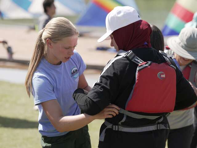 A female Summit Journeys team member helps fit a red life jacket onto a female youth participant dressed in a burgundy hijab and white hat.