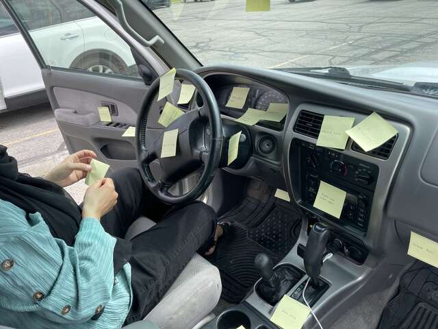 Refugee woman sits in the driver seat of a car placing sticky notes on various parts of the car interior to practice terminology related to the car.