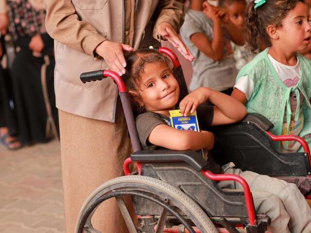 휠체어를 탄 한 어린 팔레스타인 소녀가 색연필 한 팩을 품에 안고 미소 짓고 있습니다.