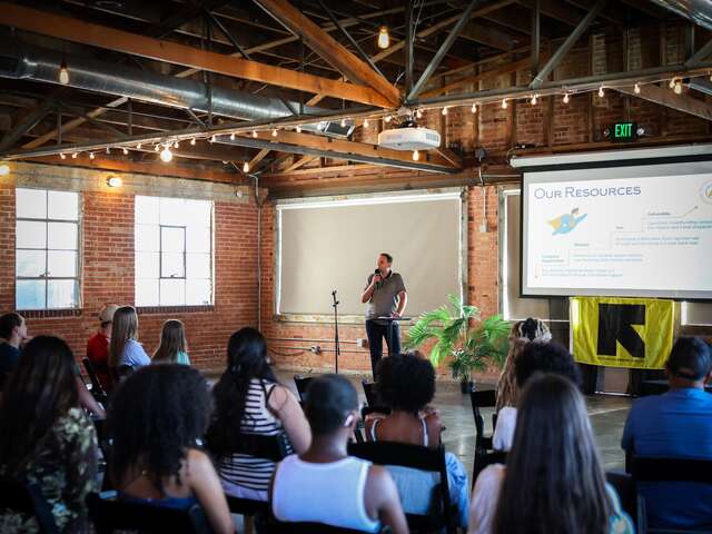 Dmytro Mateiko stands at the front of the room, back to the brick wall and projector screen, as he pitches his small business to the audience. The photo was taken from the back of the audience with numerous seated individuals in the foreground.