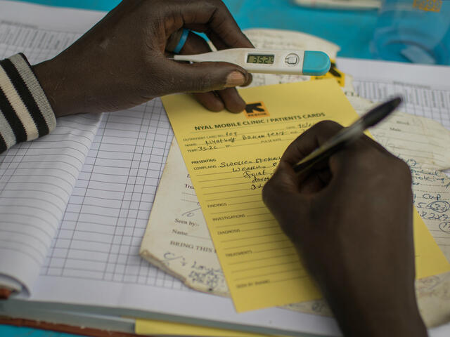 A health worker holds a thermometer indicating a patient's fever, and records the reading on a form.