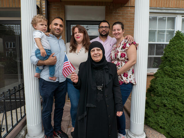 Maha and her family pose on the front porch of a house. Maha stands in front holding an American flag. Her son stands to her right holding her grandson and next to him are his wife and her other son and daughter-in-law.