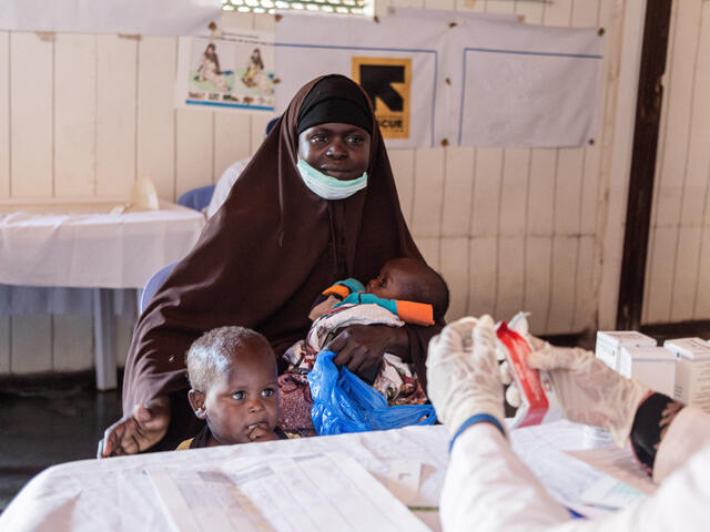 Amina sits at a table while holding a baby and putting her arm around her young son standing next to her. We can see a doctor's gloved hands holding therapeutic food.