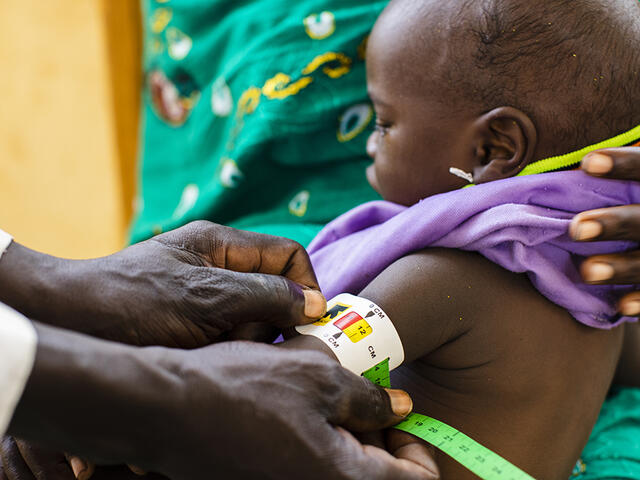 A mom holds a small child on her lap while we see the hands of a community health worker holds a MUAC (middle upper arm circumfrence) band around his arm.