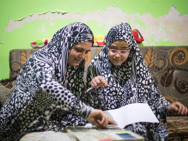 Wearing matching outfits, Ruba and Salam sit on a couch while Ruba helps her Salam with her homework.