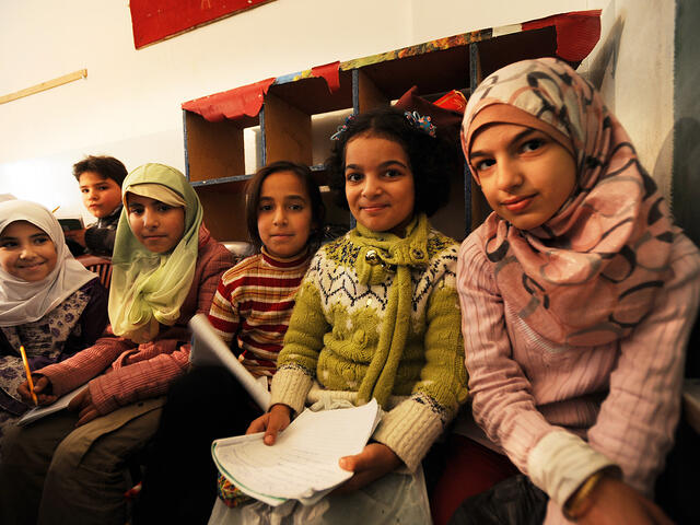 Four young girls sit next to one another in a school, looking at the camera and holding school supplies.