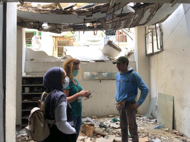 Three people stand, talking, in a damaged building. The ceiling is buckling, part of the front wall is missing and there is debris covering the ground.