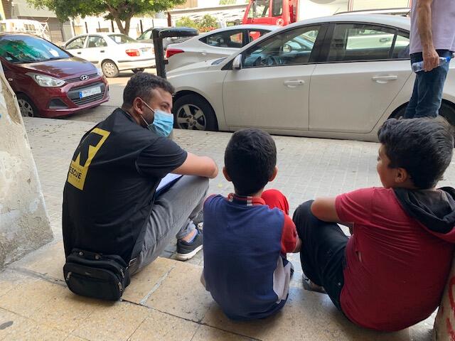 A man with an IRC vest sits on a curb with two young boys. They are all facing away from the camera and talking.