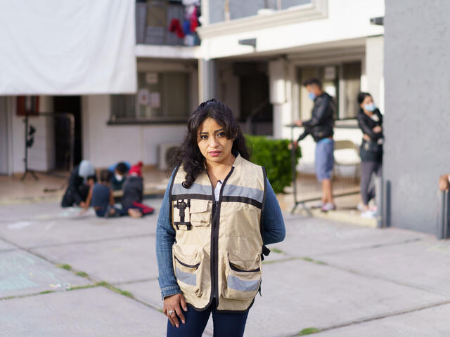Rosa, wearing a brown vest with large pockets, stands in front of the triage hotel she helps manage in Ciudad Juárez, Mexico. In the background, children draw with chalk on the ground with their parents while other adults converse.